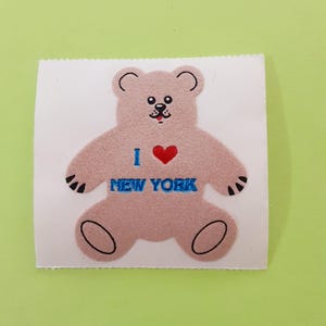 May include: A light brown teddy bear sticker with the text "I ❤️ NEW YORK". The bear has black paws, ears, and eyes, and a red heart. The sticker is on a white background.