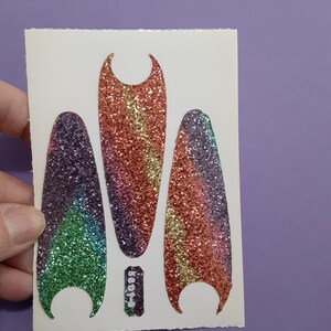 May include: A sheet of nail art stickers featuring three glittery, elongated shapes in rainbow colors, with a small rectangular piece below. The shapes are in shades of green, purple, orange, and pink. The stickers are held by a hand.