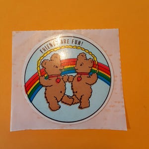 May include: A circular sticker featuring two brown teddy bears holding hands, each with a red heart. The bears stand on a rainbow with the text "FRIENDS ARE FUN!" in a yellow border against a light blue background.