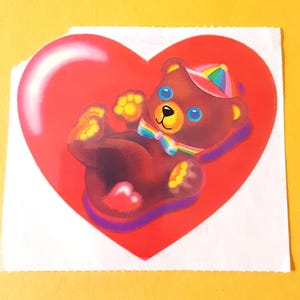 May include: A heart-shaped sticker featuring a brown teddy bear with blue eyes, wearing a rainbow-striped hat and a matching rainbow bowtie. The bear is positioned against a bright red heart background with a small pink heart on its belly.