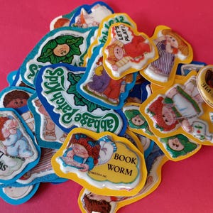 May include: A collection of vintage Cabbage Patch Kids stickers with various designs. The stickers feature illustrations of the dolls, along with text such as "Sweet Dreams," "Book Worm," and "Let's Play!" The stickers are on a pink background.