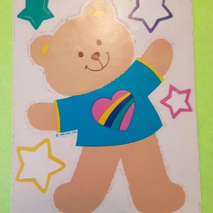 May include: A rectangular sticker sheet featuring a light brown teddy bear wearing a bright blue t-shirt with a pink heart and rainbow stripes. The bear is surrounded by four stars in teal, white, pink, and yellow. Text reads 1986 HMK. CDS.