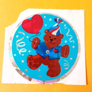 May include: A circular holographic sticker featuring a brown teddy bear wearing a blue vest and a red, white, and blue party hat. The bear holds a red heart-shaped balloon against a bright blue background with white confetti and streamers.