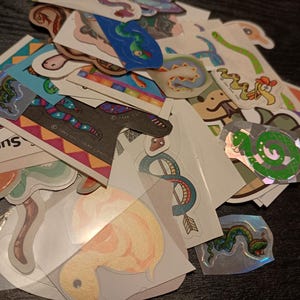 May include: A collection of colorful stickers featuring various cartoon snakes and other creatures. The stickers have a variety of designs, including a green snake, a blue snake, and a snake with a red hat. Some stickers have text.