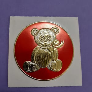 May include: A gold-colored teddy bear design on a red circular background. The bear is holding a bow and is set against a white square. The design has a 3D embossed effect.