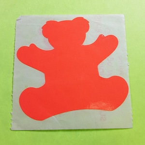 May include: A bright orange sticker in the shape of a teddy bear with outstretched arms. The sticker is on a rectangular piece of paper with a torn edge, set against a green background.