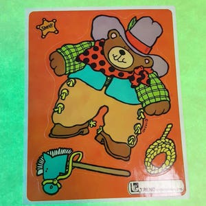 May include: A vintage sticker featuring a cartoon bear dressed as a cowboy. The bear wears a hat, scarf, and vest, with a sheriff's badge, a toy horse, and a rope. The background is orange.