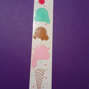 Puede incluir: Una pegatina de un cono de helado con tres bolas de helado. Las bolas son verde, marrón y rosa. El cono es marrón y blanco. Hay chispas rojas, marrones y verdes en la pegatina.