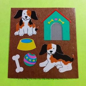 May include: A brown square with colorful dog-themed stickers. The stickers include two dogs, a dog house, a bone, a food bowl, and a ball with stars. The dogs are white, black, and orange.