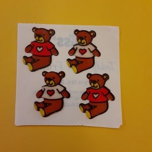 May include: Four brown teddy bear stickers with red shirts and white hearts on a white background. The stickers are on a sheet of paper.