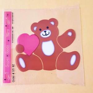 May include: A brown teddy bear sticker holding a pink heart, featuring white accents on the ears, belly, and paws. To the left, a pink vertical strip displays the text Super Toots, trumpet icons, and the label Order # ST1 RZ.