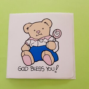 May include: A square card featuring a cartoon teddy bear holding a lollipop. The bear is tan with pink accents and a blue skirt. The text "GOD BLESS YOU!" is printed below the bear in black.