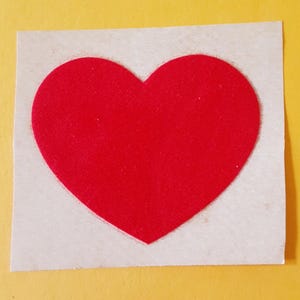 Puede incluir: Una forma de corazón de color rojo brillante recortada de un material plano, centrada sobre un cuadrado de papel de color beige claro. El corazón presenta un color rojo sólido y liso con un contorno definido y un contraste claro.