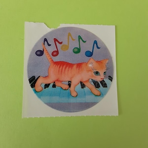 Puede incluir: Una pegatina con un gato atigrado naranja caminando sobre un teclado de piano. Notas musicales coloridas flotan sobre el gato. El gato tiene ojos azules y está sobre un fondo azul y morado.