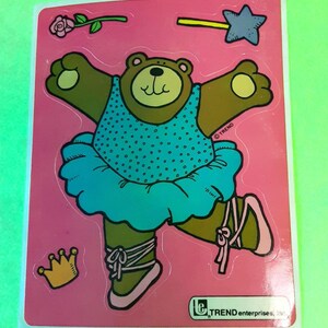 May include: A sticker featuring a cartoon bear in a blue tutu and ballet shoes. The bear has brown fur and is wearing a blue polka-dot top. A rose, star wand, and crown are also included in the design. The sticker is on a pink background.
