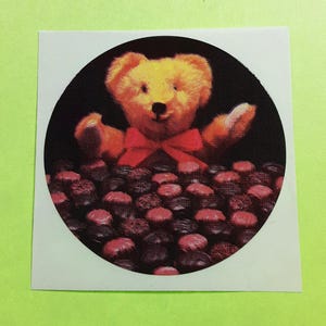 May include: A sticker featuring a teddy bear with a red bow tie, emerging from a bed of chocolates. The bear has golden fur and is set against a black background within a circular frame. The chocolates are a mix of dark and pink hues.