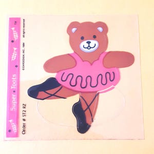 May include: A clear sticker featuring a brown teddy bear wearing a pink tutu and black ballet slippers. The sticker includes a pink vertical strip on the left side with the text Super Toots, and text reading Cardesign, Inc., 1984 and Order # ST2 RZ.