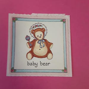 May include: A square sticker featuring a cartoon baby bear in a brown bear suit and a white bonnet with red flowers. The bear holds a blue and purple rattle. The words "baby bear" are written below the bear.