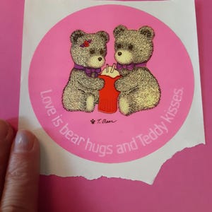 May include: A round sticker with a pink background featuring two teddy bears holding a red container. The bears have purple bows and a red flower. The text reads "Love is bear hugs and Teddy kisses."