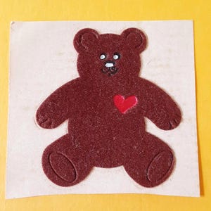 May include: A dark brown felt teddy bear applique with a textured surface, featuring white eyes, a black stitched nose and mouth, and a small, bright red heart shape centered on the chest, all set against a light beige square backing.
