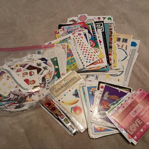 Puede incluir: Una colección de pegatinas y hojas de pegatinas de varios tamaños y diseños. Las pegatinas incluyen imágenes de estrellas, corazones y otros elementos decorativos. Algunas pegatinas están empaquetadas en bolsas y hojas de plástico transparente.