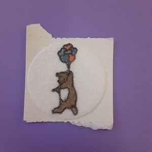 May include: A brown bear illustration holding colorful balloons, set on a white circular patch. The patch is affixed to a torn piece of paper, all against a purple background. The bear is drawn in a simple, whimsical style.