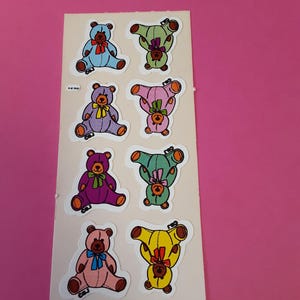 May include: Sheet of colorful teddy bear stickers. Each bear has a different color, including blue, green, purple, pink, and yellow. The bears are sitting or upside down, with bows around their necks. The stickers are on a white background.