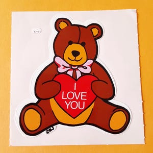 May include: A brown teddy bear sticker with a light brown muzzle and inner ears, wearing a light pink bow around its neck. The bear holds a large red heart featuring the white text I LOVE YOU. The sticker has a white border and a small BJ logo.