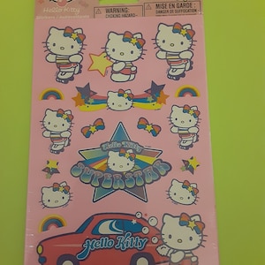 Puede incluir: Una hoja rosa de pegatinas con Hello Kitty en varias poses. Las pegatinas incluyen a Hello Kitty en patines, un coche y una estrella con las palabras "Hello Kitty Superstar".