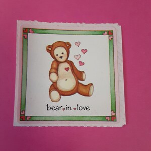 May include: A square sticker with a brown teddy bear illustration. The bear has a heart on its chest and is surrounded by pink hearts. The text "bear in love" is below the bear. The sticker has a green border with pink hearts.