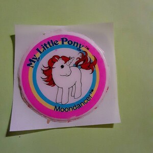 May include: A pink and blue circular sticker with the text "My Little Pony" and "Moondancer" in a white circle with a rainbow border. A white unicorn with a red mane and tail is in the center of the circle.
