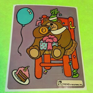 May include: A colorful sticker featuring a cartoon bear wearing a party hat and sitting on a chair. The bear holds a party horn and a gift box. A blue balloon and a slice of cake with a lit candle are also depicted.