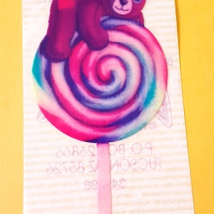May include: A rectangular sticker featuring a purple bear with a pink bow on its head, resting on a large, round, multi-colored swirl lollipop with pink, blue, and purple hues. The sticker has a light-colored background with faint text.