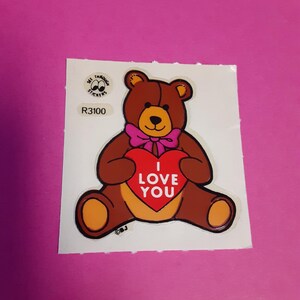 May include: A brown teddy bear sticker with a pink bow tie and a red heart that says "I LOVE YOU". The sticker has a white background and the text "R3100".