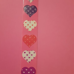 Puede incluir: Una tira de pegatinas en forma de corazón en varios colores y patrones. Las pegatinas incluyen corazones blancos con diseños florales azules, corazones morados con diseños florales blancos, corazones blancos con lunares rojos, corazones rojos sin patrón, corazones blancos con corazones morados.