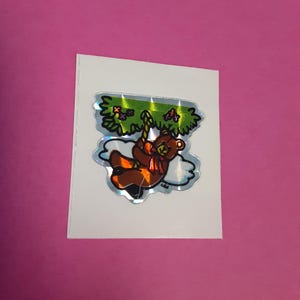 May include: A holographic sticker featuring a cartoon bear swinging from a tree branch. The bear is brown with orange accents and a red scarf. The tree has green leaves and the sticker has a white border.
