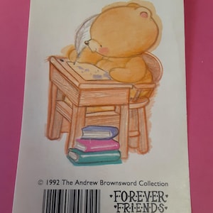 May include: A Forever Friends card featuring a cartoon bear writing at a small wooden desk. The bear is light brown with a pink nose and is seated on a matching chair. A stack of three books in blue, pink, and teal are at the base of the desk. The card has text and a barcode.