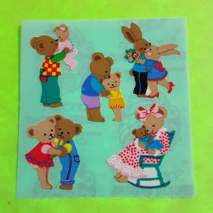May include: A sheet of colorful stickers featuring various cartoon bears and a bunny hugging each other. The bears are dressed in different outfits, including dresses, overalls, and sweaters. The background is a light green color.