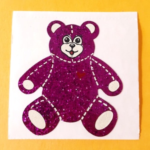 May include: A purple glittery teddy bear cutout featuring white stitched detailing around the limbs and ears, a white muzzle with a black nose and mouth, and a small red heart accent on the chest, all centered on a plain white square background.