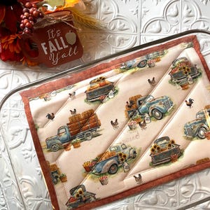 May include: A quilted hot pad with a fall theme featuring a vintage truck carrying hay bales and pumpkins. The hot pad is beige with a brown border and a repeating pattern of trucks, hay bales, pumpkins, and sunflowers. The text "It's Fall Y'all" is visible in the background.