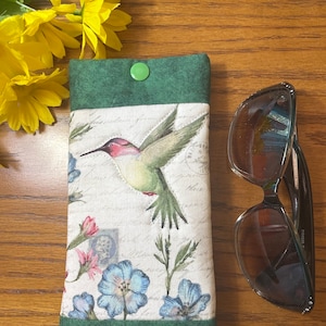 May include: Green fabric eyeglasses case with a hummingbird and floral print. The case has a green button closure.