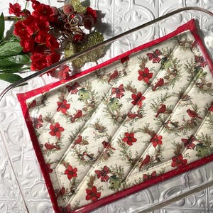 May include: A red and white quilted hot pad with a Christmas bird and floral pattern. The hot pad is placed on top of a clear glass baking dish.
