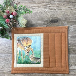 May include: A brown quilted pot holder with a rectangular image of a fawn and a rabbit in a field. The pot holder has a light beige border around the image and a vertical stitched pattern on the right side.