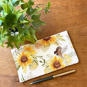 May include: A white fabric pencil case with a sunflower and bird print. The case is lying on a wooden surface with a pen next to it.
