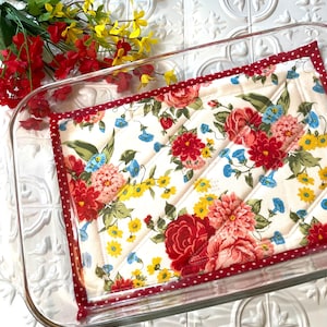 May include: A clear glass baking dish with a floral patterned quilted pot holder. The pot holder features a white background with red, pink, blue, and yellow flowers. The edge of the pot holder is red with white polka dots.