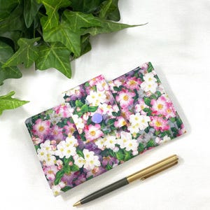 May include: A floral print wallet with a purple snap closure. The wallet features a pattern of pink, white, and purple flowers with green leaves. A gold and black pen rests beside the wallet on a white surface.