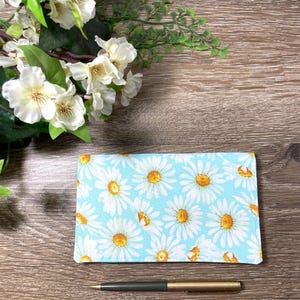 May include: A light blue fabric wallet with a white daisy pattern and a gold and black pen on a wooden surface. White flowers and greenery are in the background.