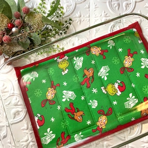 May include: A green fabric pot holder with a red border. The fabric has a repeating pattern of cartoon characters from the movie "How the Grinch Stole Christmas!" The characters include the Grinch, Cindy Lou Who, and Max the dog. The pot holder is in a clear glass baking dish.