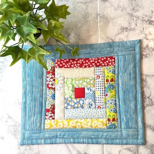May include: A blue quilted square with a log cabin pattern in red, yellow, white, and green fabrics. The quilt is made of various floral prints and has a blue border.