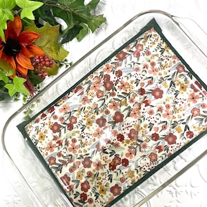 May include: A rectangular glass baking dish containing a fabric cover with a floral pattern featuring small red, orange, and yellow flowers on a cream background, finished with a dark green border trim around the edges of the fabric.